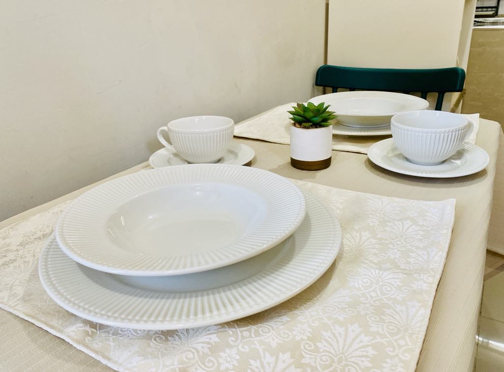 A dining table is set with white dishware, including plates and cups arranged neatly. A small green plant is positioned at the center, surrounded by placemats that offer a subtle textured pattern, complementing the light-colored tablecloth.