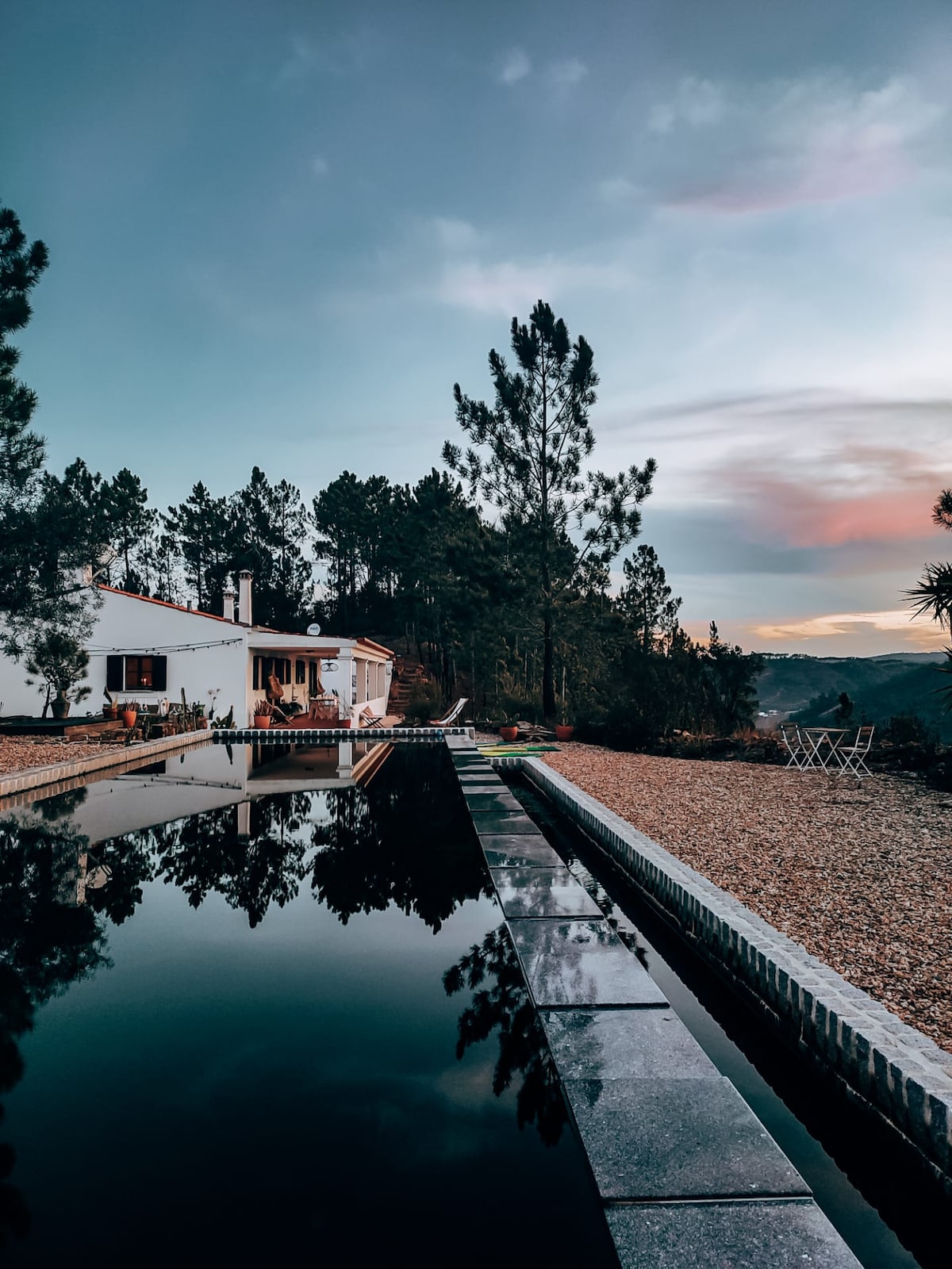A tranquil natural pool reflects the surrounding trees and sky, bordered by a stone pathway. The white house is visible in the background, along with outdoor seating. The scene is complemented by soft colors of dusk.