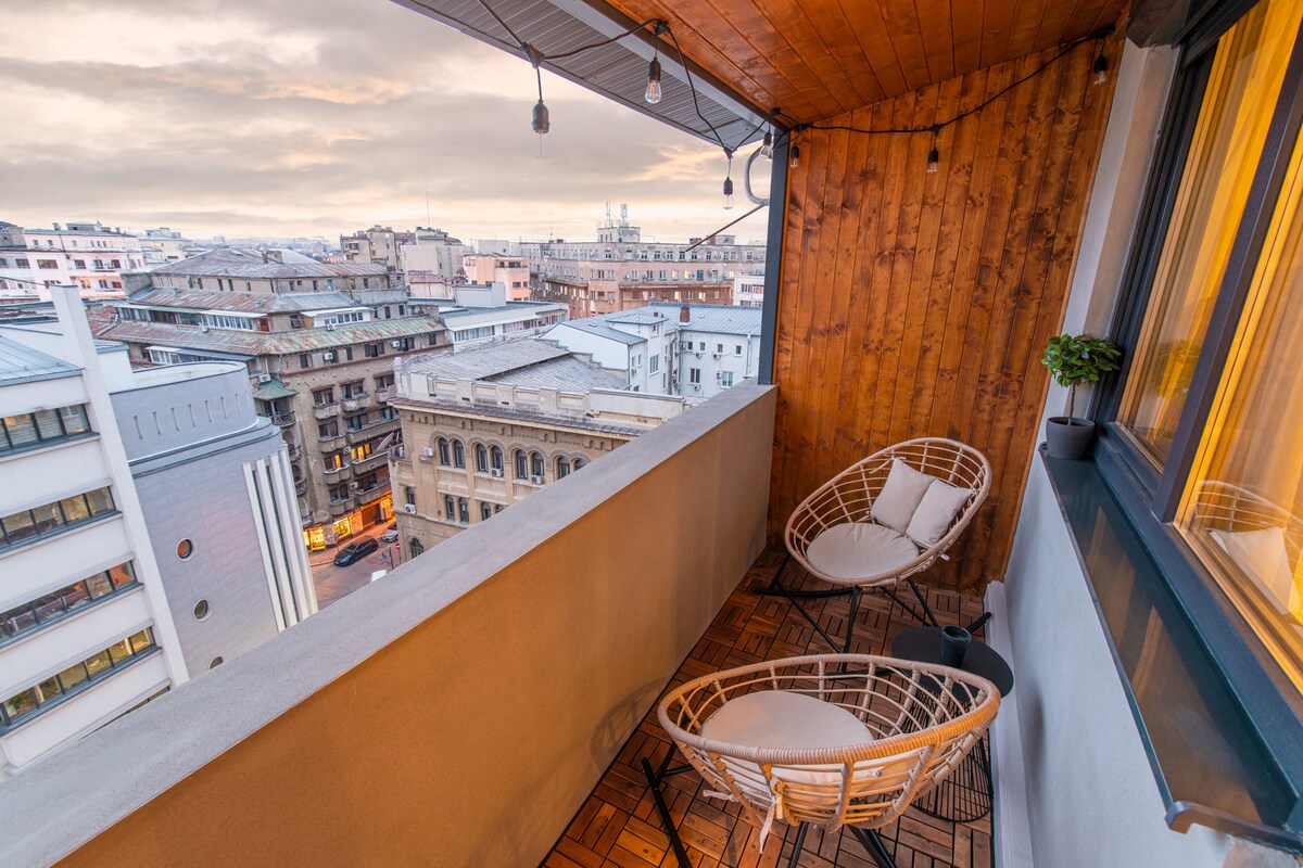 A private terrace is featured, offering a view of the cityscape. Two stylish chairs are positioned on a decorative wooden floor, complemented by a small potted plant. Soft lighting enhances the space, creating a suitable environment for relaxation.