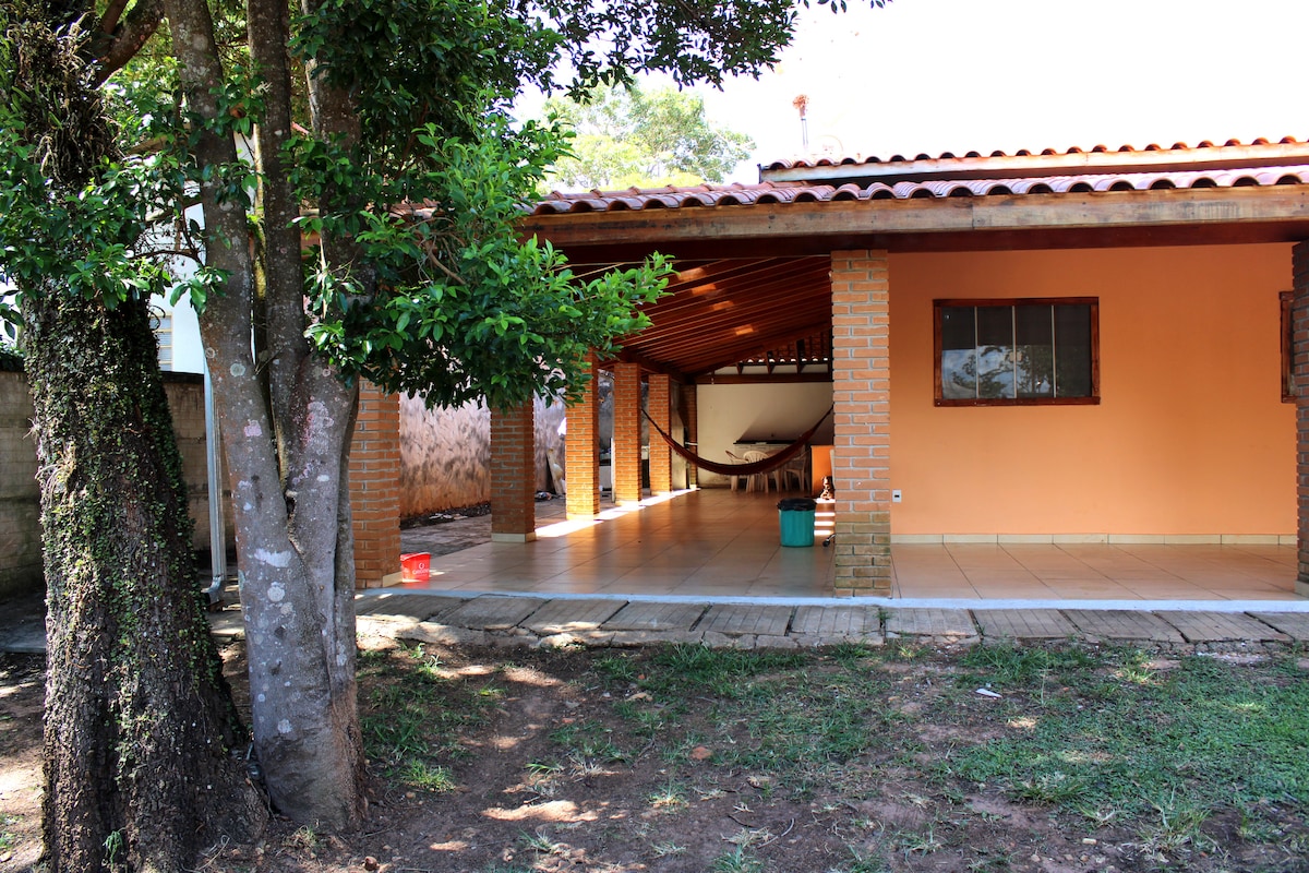 An outdoor area is presented, featuring a spacious covered patio supported by brick columns. A hammock is visible in the corner, and a tiled floor extends throughout the space. Surrounding greenery adds a natural touch, enhancing the connection with the outdoors.