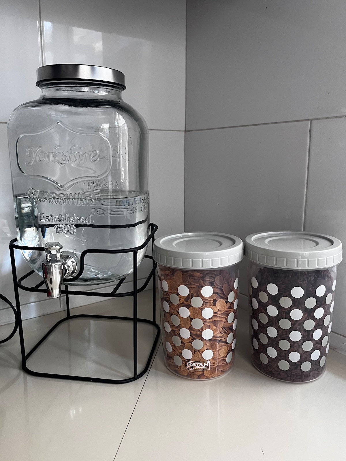A glass water dispenser is positioned on a metal stand, featuring a spout for easy access. Two containers with white polka dots sit beside it, filled with dry goods. The scene is set against a light tiled background.