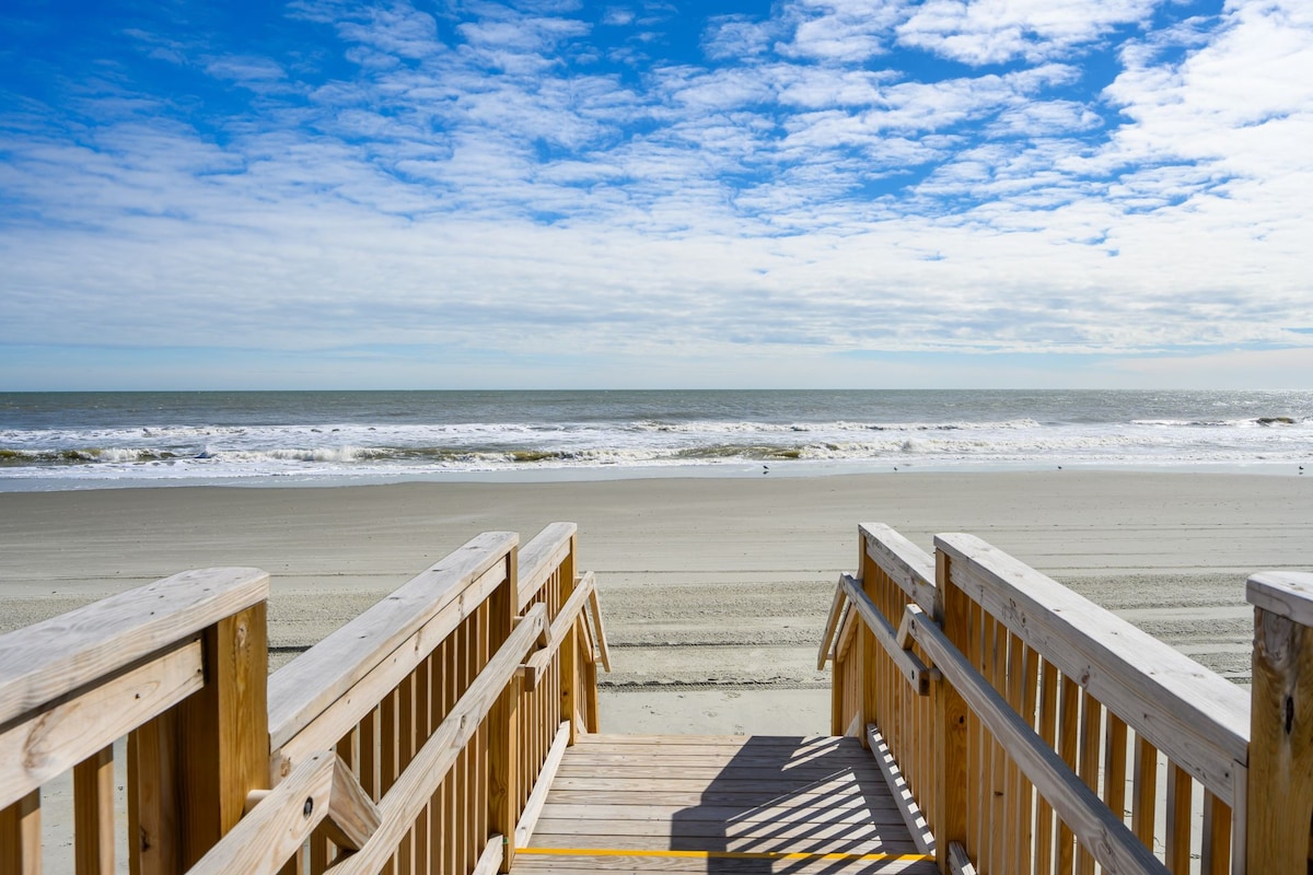 Wooden steps lead down to a wide sandy beach, with gentle waves lapping at the shore. The sky is filled with soft, fluffy clouds, allowing sunlight to filter down onto the tranquil scene. The horizon is unobstructed, creating a peaceful coastal atmosphere.