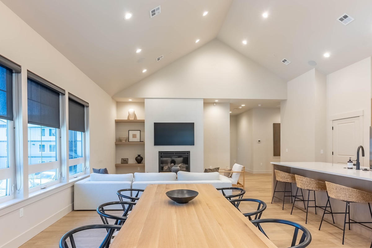 A spacious living area is illuminated by recessed lighting, featuring a large sofa and a modern television mounted on the wall. A dining table with light wood finishes is centered in the room, surrounded by black and natural-colored chairs, with large windows allowing natural light to enter.