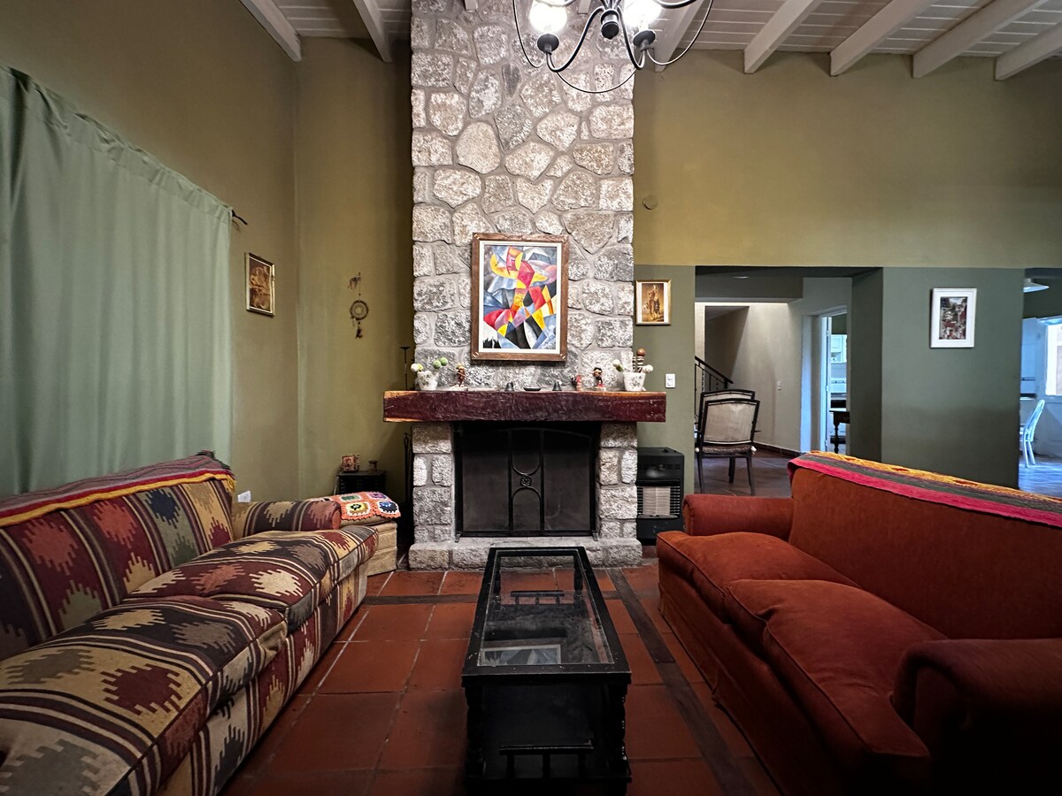 A living room features a stone fireplace as a focal point, surrounded by comfortable sofas in warm tones. A dark coffee table sits in the center, and artwork is displayed on the walls. Natural light filters through a window covered by soft drapes.