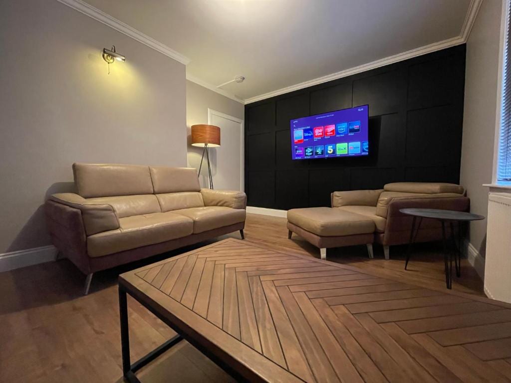 A modern living space features a light-colored leather sofa and a matching lounge chair positioned around a dark wooden coffee table. A large flat-screen TV is mounted on a black wall, while a standing lamp provides soft illumination in the corner.