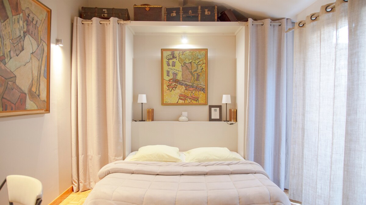 A cozy sleeping area is presented, featuring a spacious bed adorned with soft linens. Artwork displayed on the wall adds character, while two bedside lamps provide gentle illumination. Natural light fills the room through partially open, light-colored curtains.