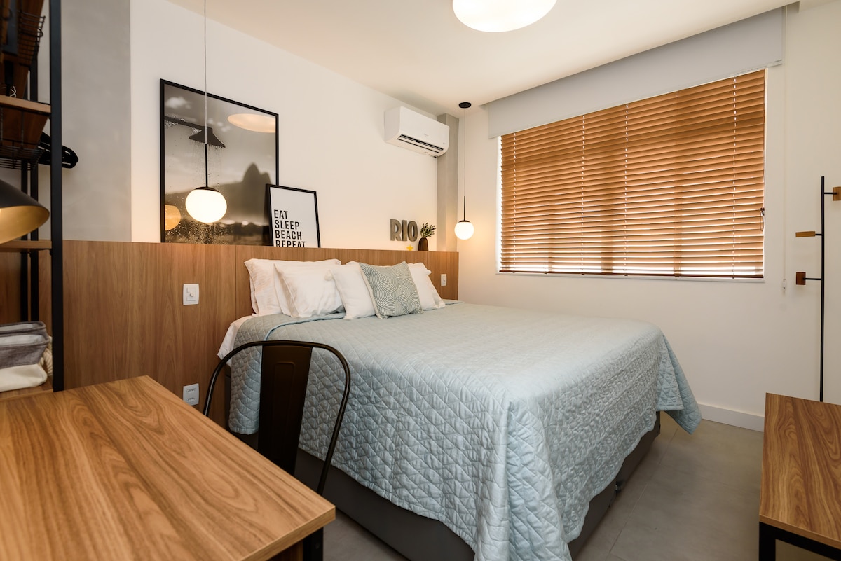 A spacious bedroom features a king-size bed dressed in light teal bedding. Natural light filters through the wooden blinds, illuminating the contemporary decor which includes framed art and a small desk. A wall-mounted air conditioning unit provides comfort.