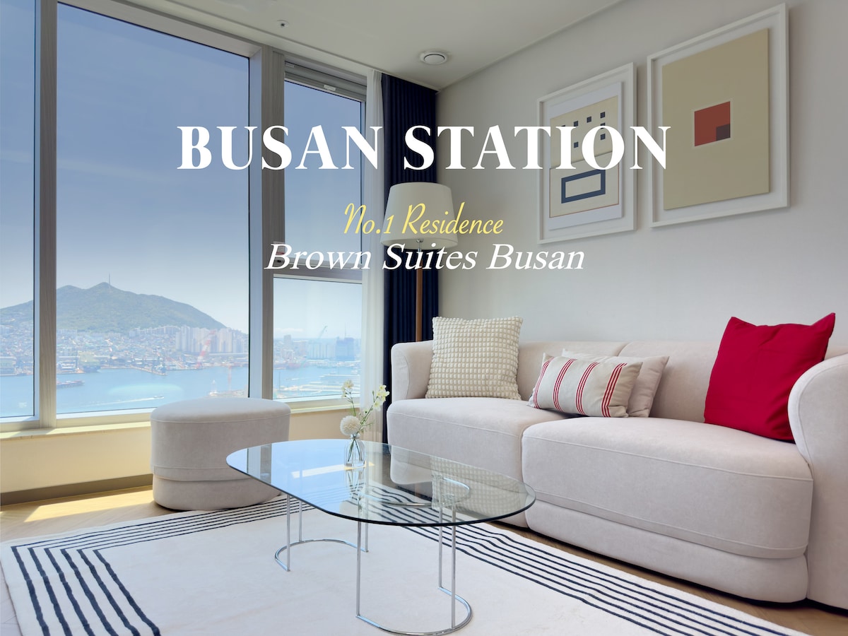 A well-lit living space is adorned with a comfortable sofa, accented by colorful cushions. A sleek glass coffee table is positioned in front of the sofa. Large windows provide a view of the ocean and cityscape, enhancing the atmosphere with natural light.