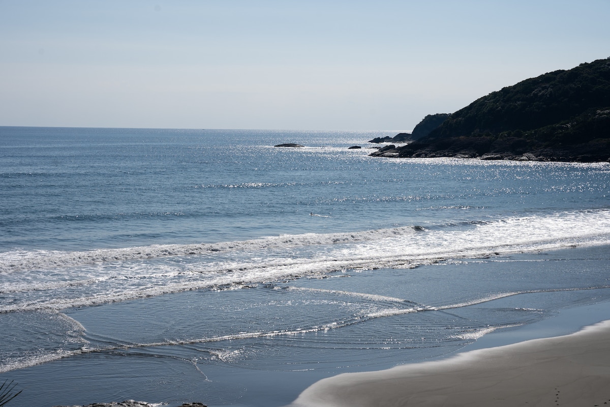 A serene view of the ocean is captured, showcasing gentle waves rolling onto a sandy beach. The water reflects sunlight, creating a shimmering effect. A distant coastline with lush greenery can be seen, complementing the tranquil atmosphere.