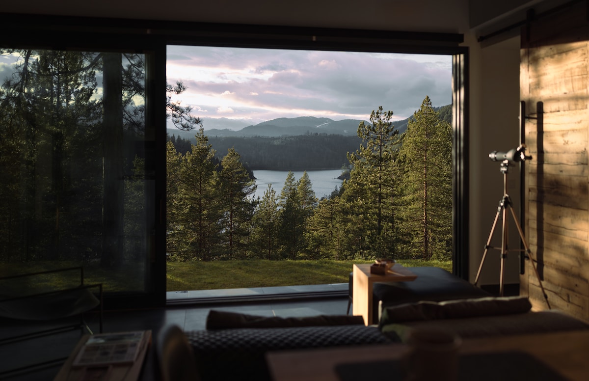A large sliding door opens to reveal a scenic view of a lake surrounded by trees and mountains. Inside, natural light fills the space, highlighting a telescope positioned nearby and wooden accents throughout the room.