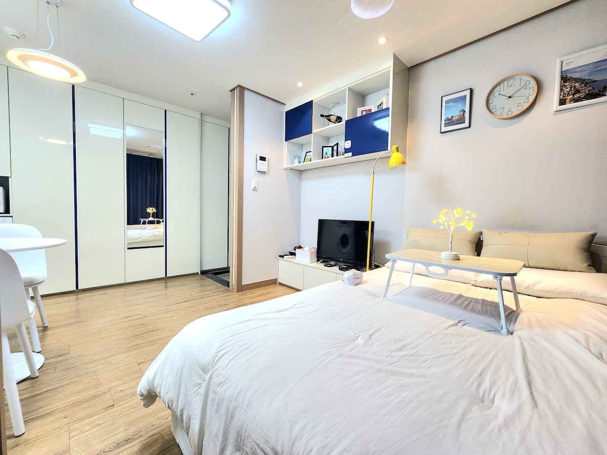A spacious room features a comfortable bed with soft linens. A modern TV is positioned on a unit beneath a shelf displaying decorative items. A small dining table with chairs is visible, alongside a bright yellow lamp, enhancing the overall lighting.