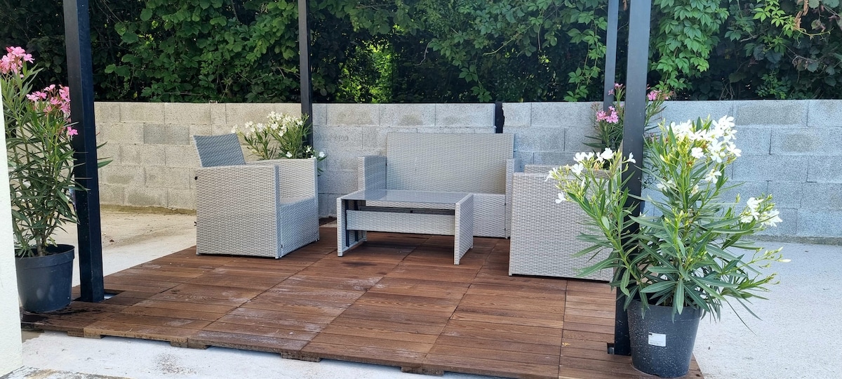 A shaded outdoor seating area features a light gray wicker couch and two chairs arranged around a rectangular coffee table. Potted plants with white flowers and dark greenery are positioned on either side of the seating, enhancing the peaceful atmosphere.