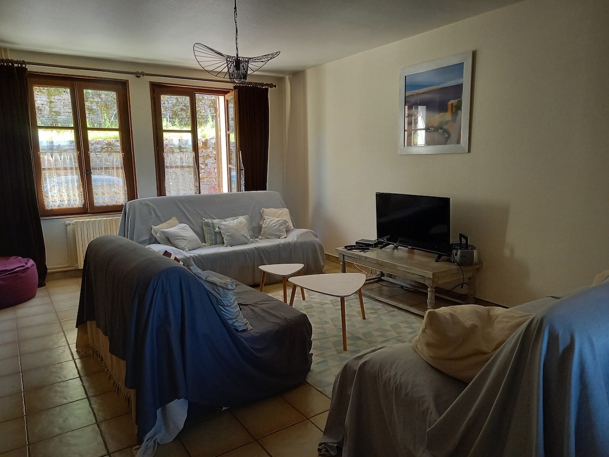 A living area features three sofas draped with light covers, arranged around a small, round coffee table. Natural light enters through two large windows, framed by dark curtains. A television is positioned on a low console, with a piece of artwork mounted on the wall.
