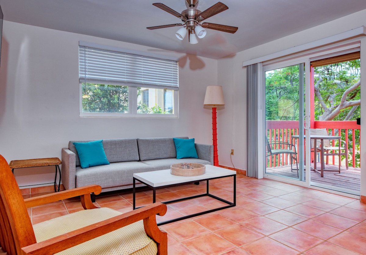 A bright sitting area is furnished with a light grey couch accented by blue cushions. A wooden chair and a contemporary coffee table are also present. Large glass doors open to a furnished terrace overlooking greenery, while a ceiling fan provides ventilation.