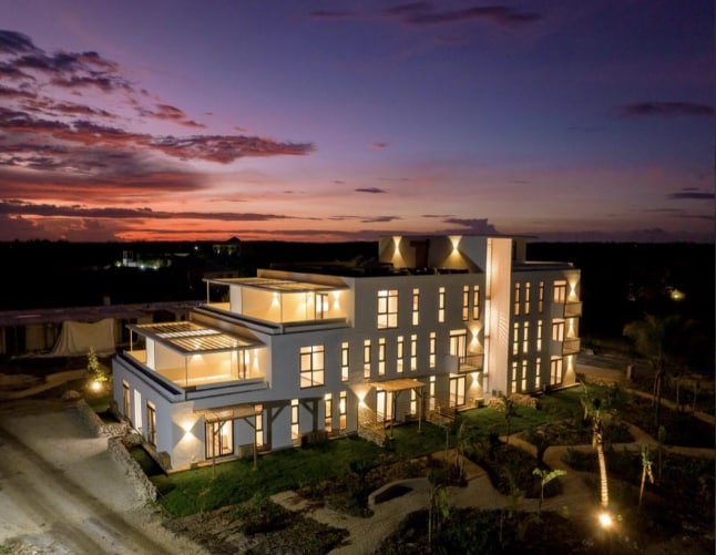 A modern two-story building is captured at dusk, with warm lights glowing from large windows. The structure features multiple balconies and a flat roof, surrounded by landscaped greenery. The sky is painted with hues of purple, orange, and pink, providing a stunning backdrop.