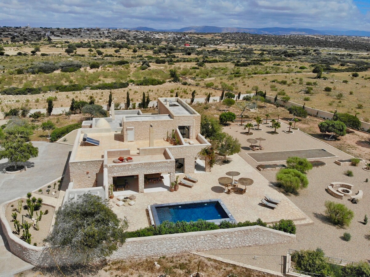 The aerial view captures a Moroccan traditional stone house surrounded by a wild garden. A rectangular outdoor pool is visible, with seating areas arranged among greenery. The vast landscape extends in the background, providing a sense of tranquility and connection to nature.