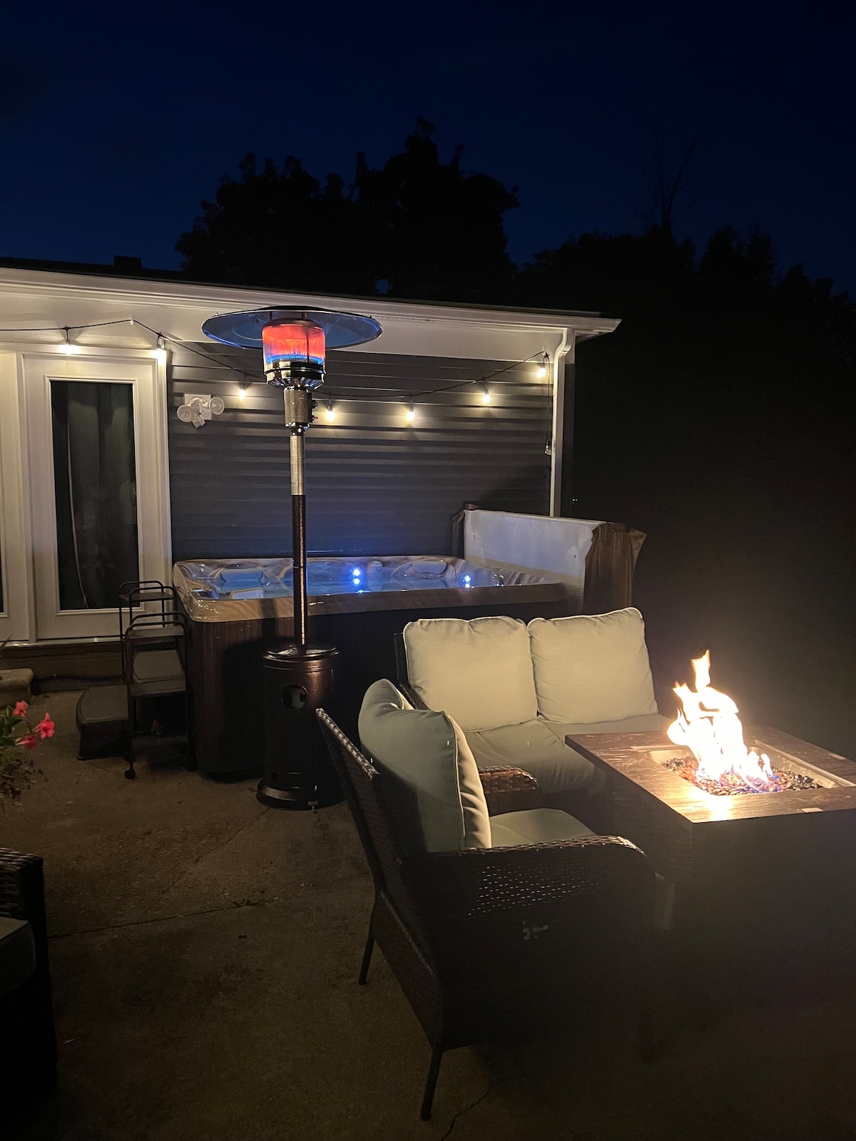 A serene outdoor space is illuminated by soft lighting, featuring a hot tub and comfortable seating area. A fire table with a flickering flame is positioned next to plush seating, creating a cozy atmosphere for relaxation under the night sky.