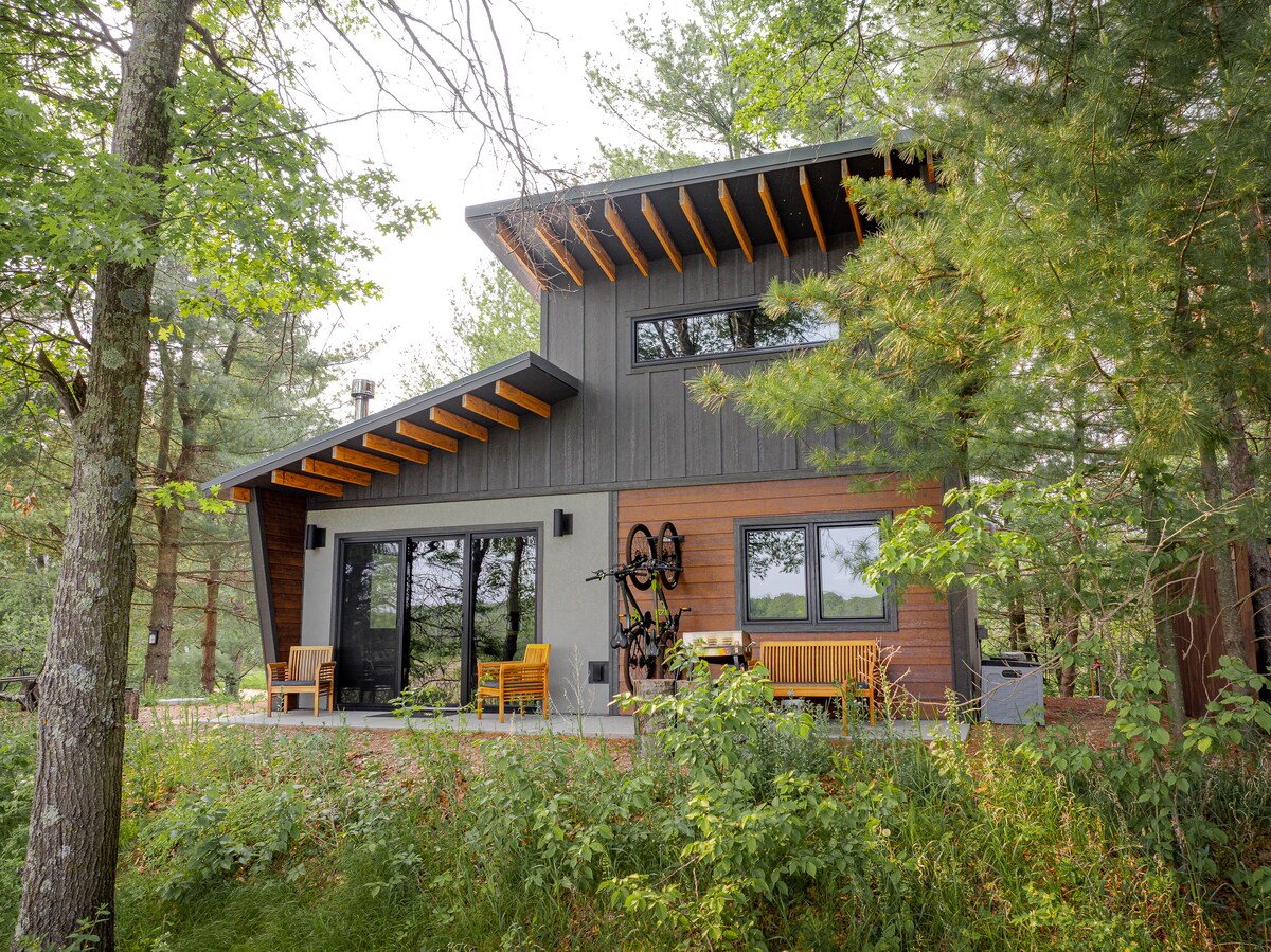 A modern cabin is set amidst trees, featuring a spacious patio with seating options. Large windows provide views of the surrounding nature, while a bicycle is mounted on the wall. The roof showcases a unique angular design, harmonizing with the natural landscape.