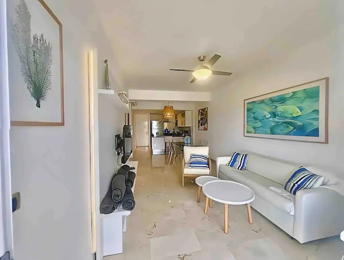 A bright living area is featured, with a white sofa and decorative cushions. A round coffee table sits in the center, while a fan overhead provides airflow. In the background, the kitchen area is visible, along with a dining space. A large wall art depicting fish adds color to the room.