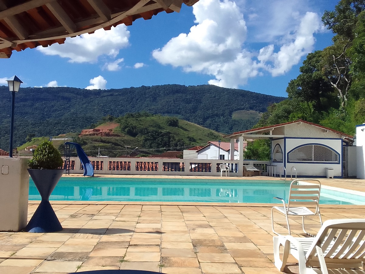 A clear blue swimming pool is featured in the foreground, surrounded by sun loungers and a light-colored tiled patio. The backdrop includes rolling green hills under a partly cloudy sky, adding a natural ambiance to the space.