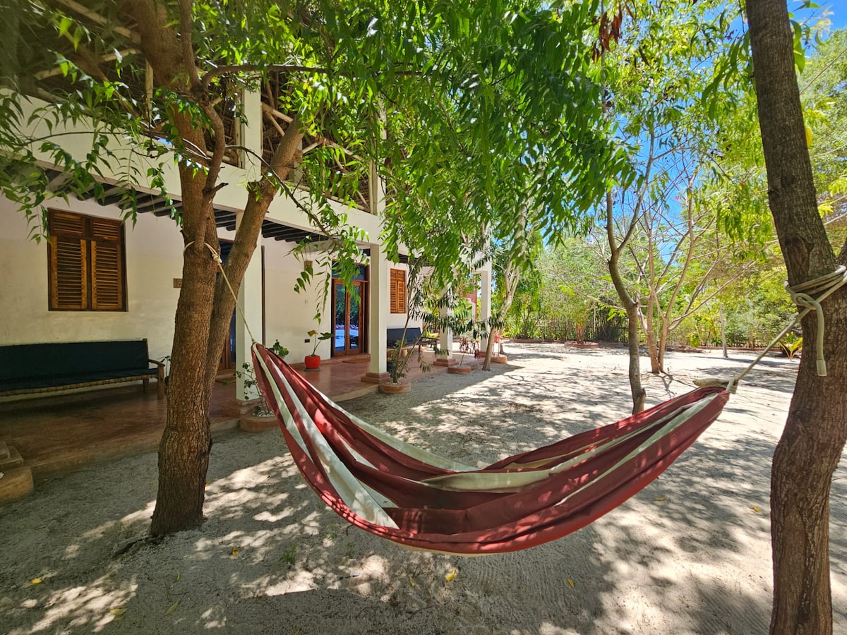 A spacious garden area is depicted, with a hammock strung between trees, shaded by lush greenery. The ground is covered in soft sand, and a nearby bench can be seen, complementing the tranquil atmosphere of the surrounding space.