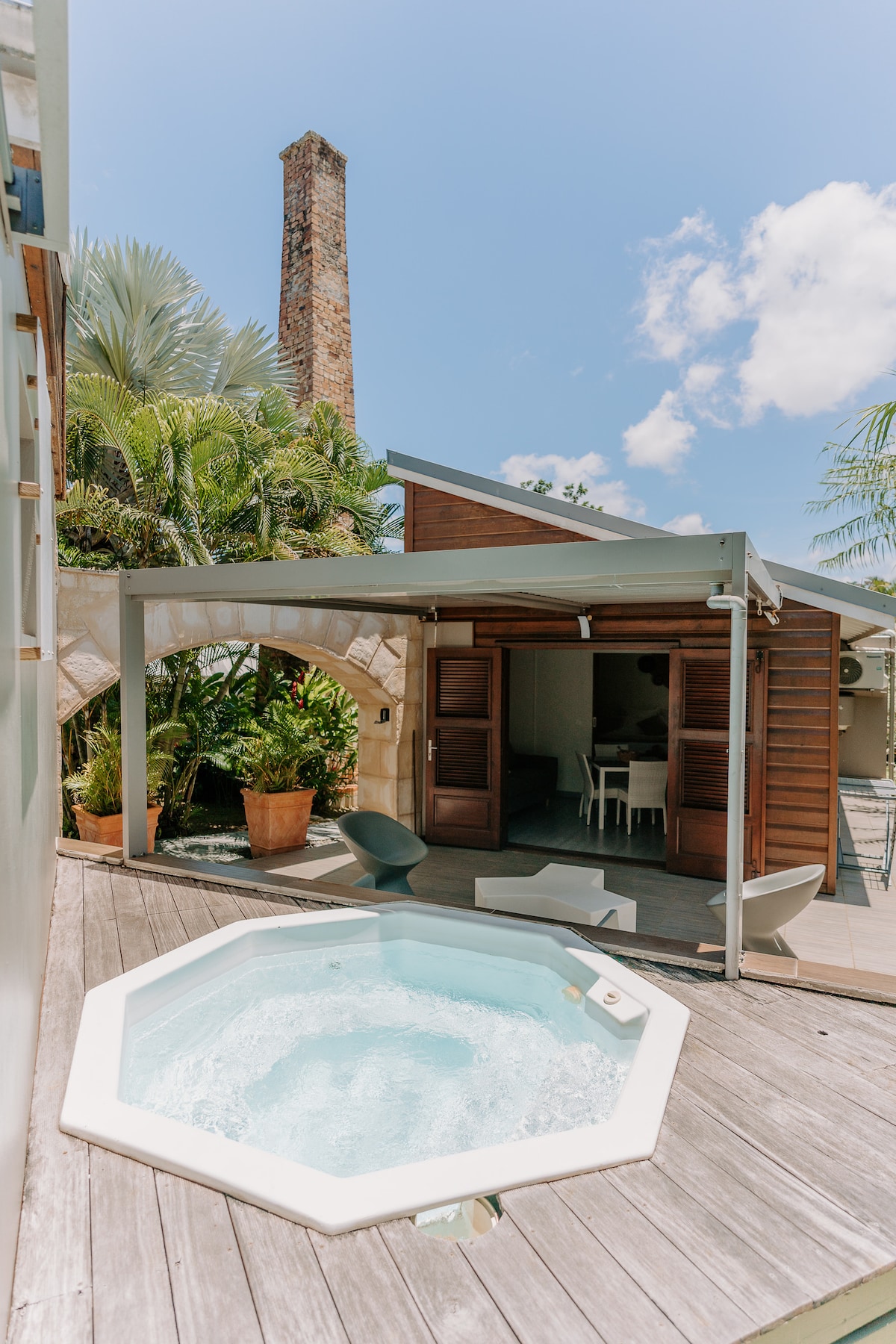 An outdoor area features a private octagonal pool surrounded by wooden decking. Lush tropical plants provide a natural backdrop, while a charming brick chimney adds character. A shaded seating area and a table are visible in the distance, contributing to a relaxing ambiance.