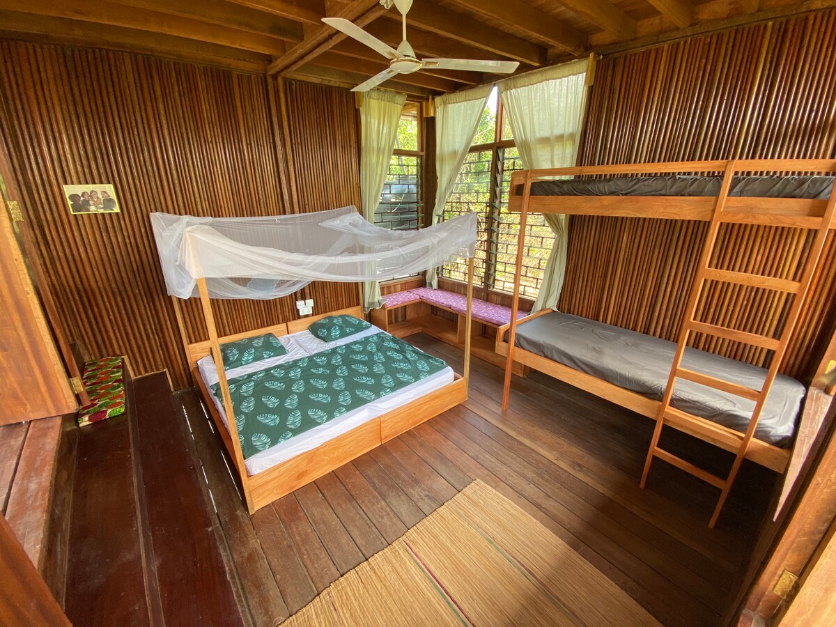 A cozy sleeping area features a spacious bed covered with a patterned green bedspread and a bunk bed positioned against a wooden wall. Natural light filters in through large windows, while a seating area with cushions offers a relaxing spot to enjoy the view.