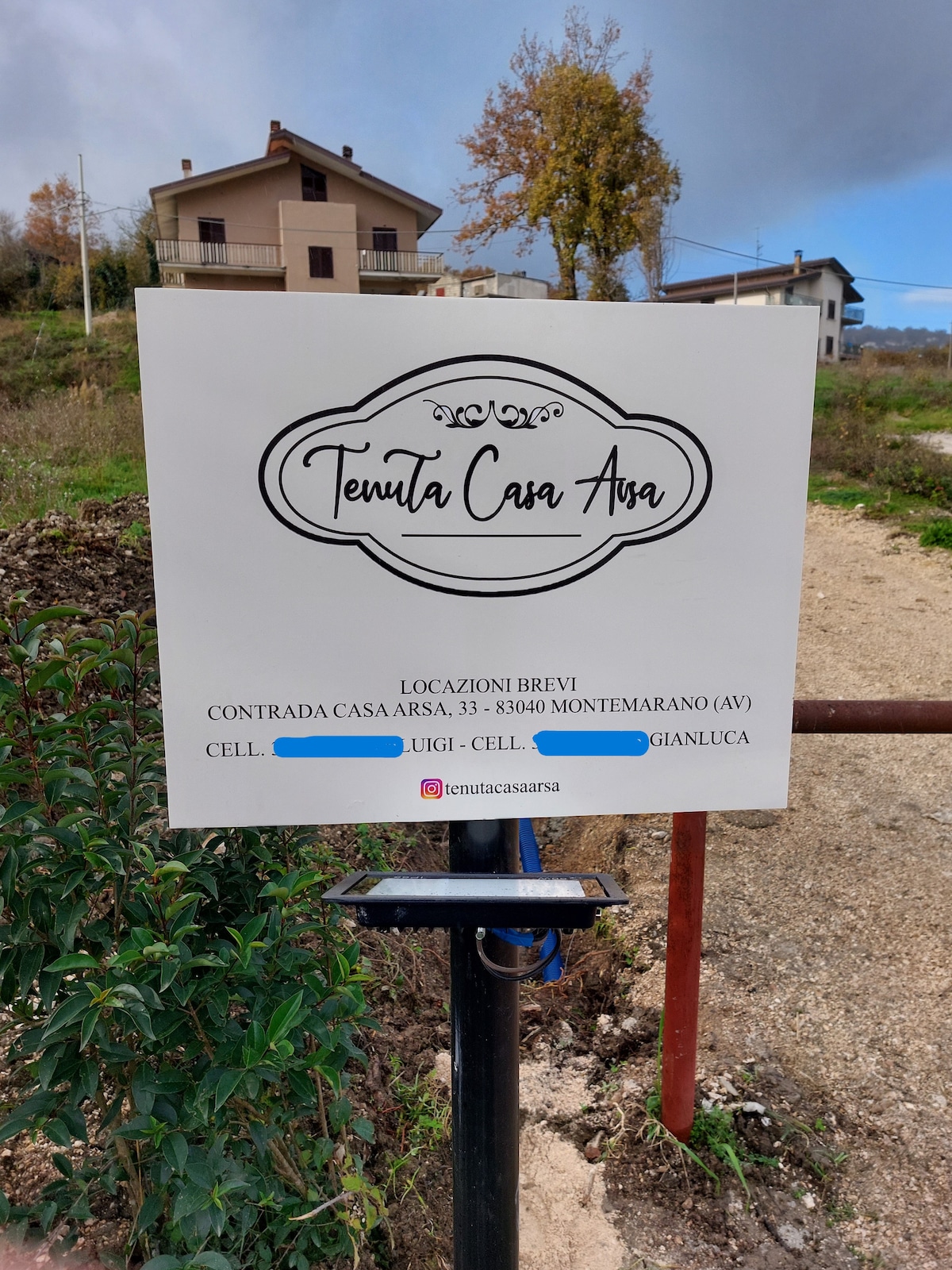 A sign for Tenuta Casa Arsa is displayed prominently, featuring the property name and location details. The background shows a dirt path leading to a villa and surrounding greenery, providing a glimpse of the natural landscape and inviting visitors to explore the area.