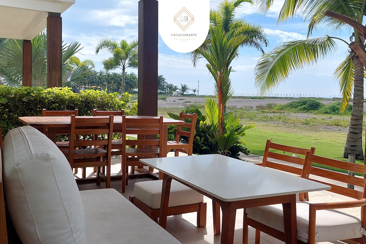 An outdoor dining area features a light wood table surrounded by six chairs, with a plush sofa positioned nearby. Lush greenery and palm trees frame the space, providing a relaxing view of the sea in the background under a bright sky.
