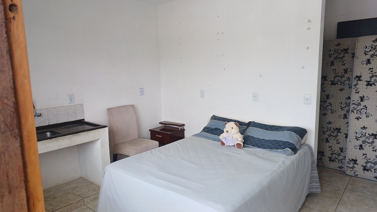 A spacious bedroom features a large bed with a light-colored coverlet and a teddy bear placed on top. A wooden nightstand and a chair are included, alongside a small kitchenette with simple countertops. Neutral walls enhance the room's calm environment.