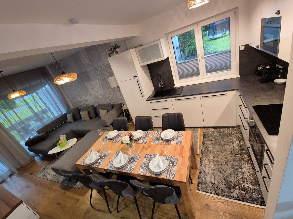 A modern kitchen and dining area are showcased, featuring sleek cabinetry and a spacious table set with white dinnerware. Large windows provide natural light, highlighting the comfortable seating arrangement and elegant decor throughout the space.