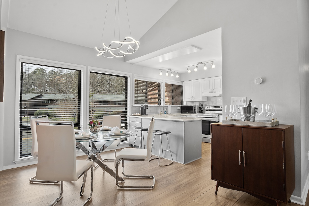 A modern dining area features a glass table surrounded by four sleek white chairs. Natural light enters through large windows with wooden blinds, illuminating the open kitchen equipped with stainless steel appliances and an island. A sideboard provides additional storage space.