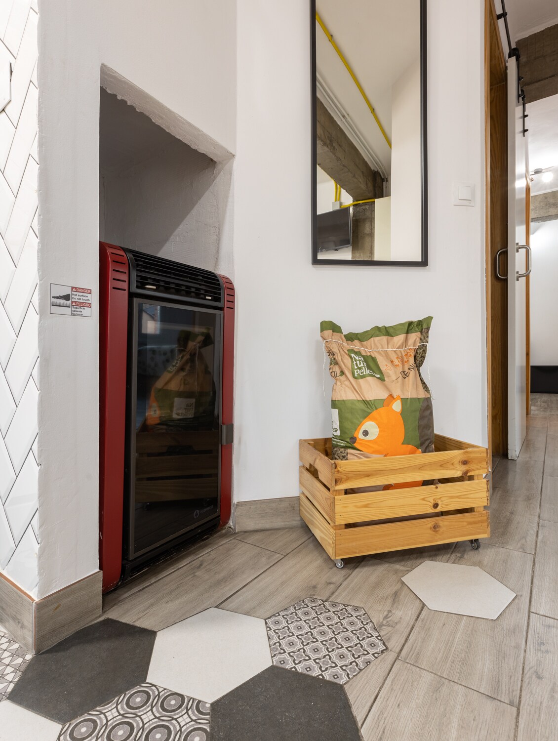 An entryway features a red heater against a white wall, complemented by a large mirror above. A wooden crate on wheels holds a colorful cushion with an orange fish design, adding a playful touch to the space. Distinctive flooring tiles in varying shapes provide texture.