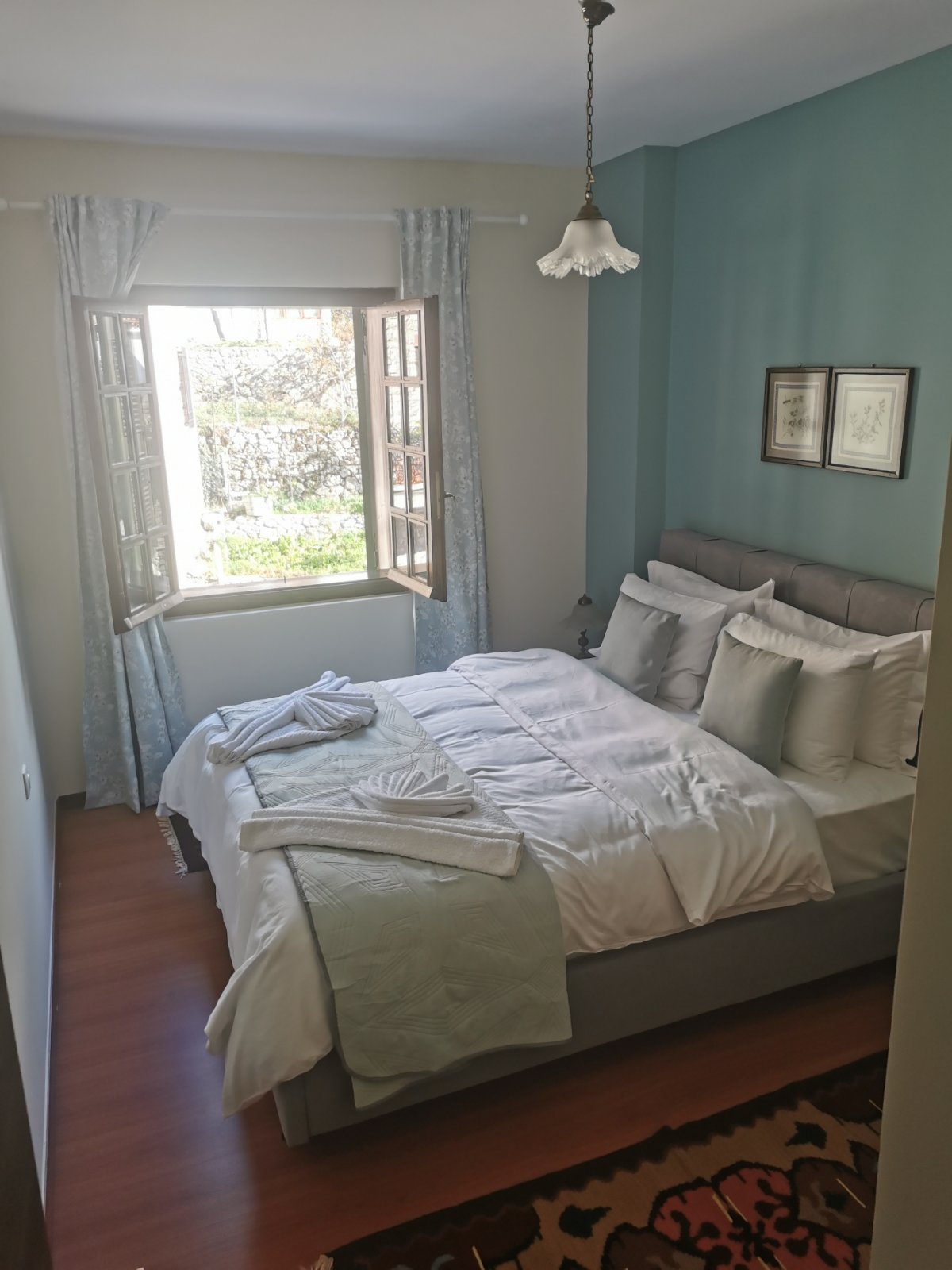 A cozy bedroom is presented with a spacious bed adorned with soft white linens and decorative pillows. Light filters through an open window flanked by light blue curtains, revealing greenery outside. A patterned rug adds warmth to the wooden floor.