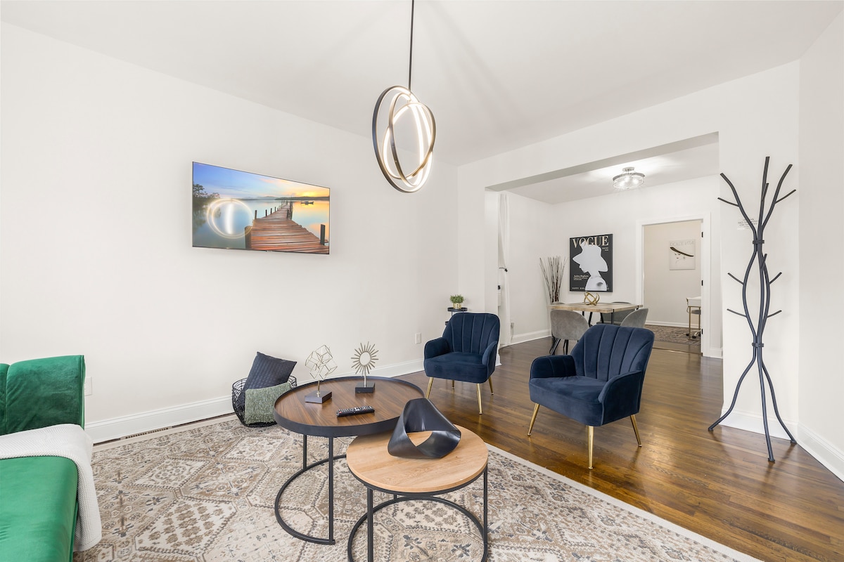 A stylish living area features a large wall-mounted TV displaying a serene landscape. Two navy blue armchairs are positioned near a wooden coffee table, which holds decorative items. A patterned area rug covers the wooden floor, while a unique floor lamp adds character to the space.