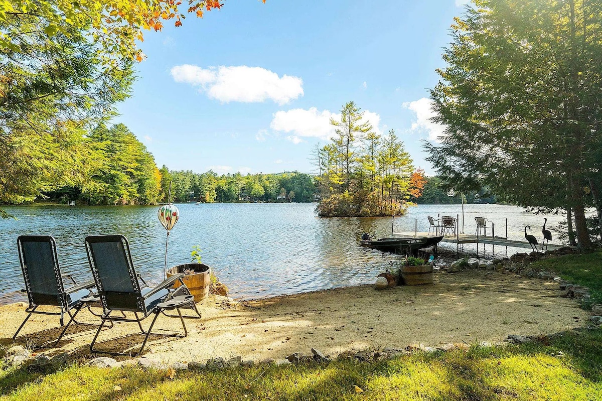 A peaceful lakeside view showcases a sandy beach area with two reclining chairs. A private dock extends into the water, surrounded by trees displaying autumn colors, while gentle waves reflect the clear blue sky.
