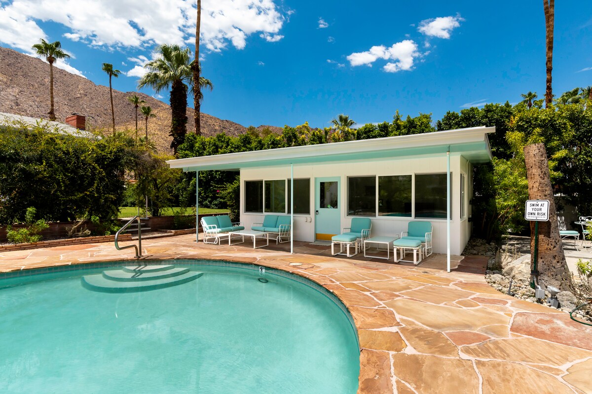 A inviting pool area features a clear turquoise pool surrounded by a stone deck. Lounge chairs line the edge of the pool, and a bright, modern poolside room with large windows is visible, providing ample views of the landscaped garden and mountain backdrop.