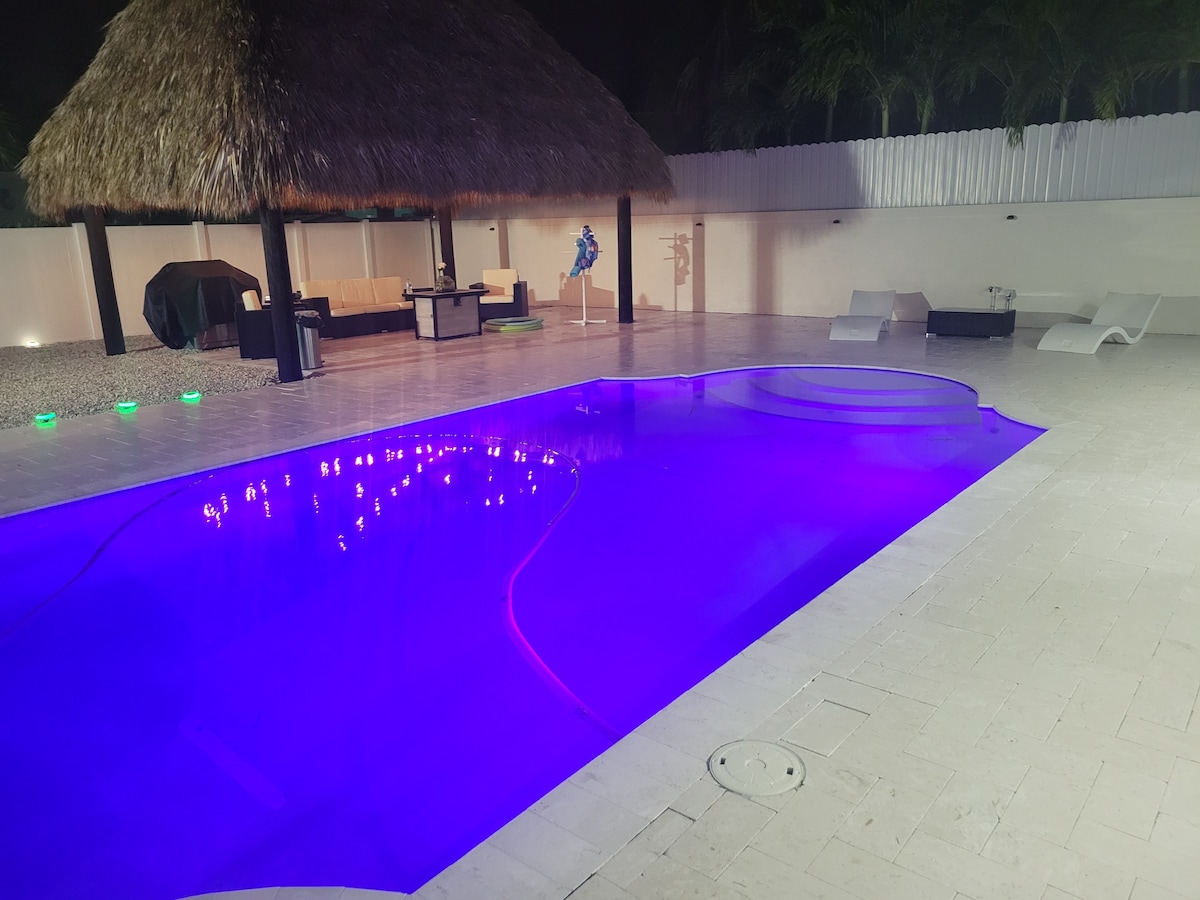 A spacious outdoor area features a well-lit pool with a calming blue glow. A thatched-roof gazebo provides shade over seating, while lounge chairs are arranged around the poolside. The surrounding area is dressed in neat concrete and enhanced by subtle landscaping.