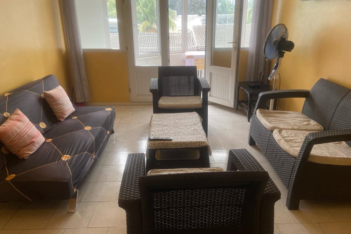 A welcoming living area features a mix of seating options, including a cozy futon and wicker armchairs. A coffee table occupies the center, while natural light filters in through large windows. Soft cushions add comfort to the seating arrangement against warm yellow walls.