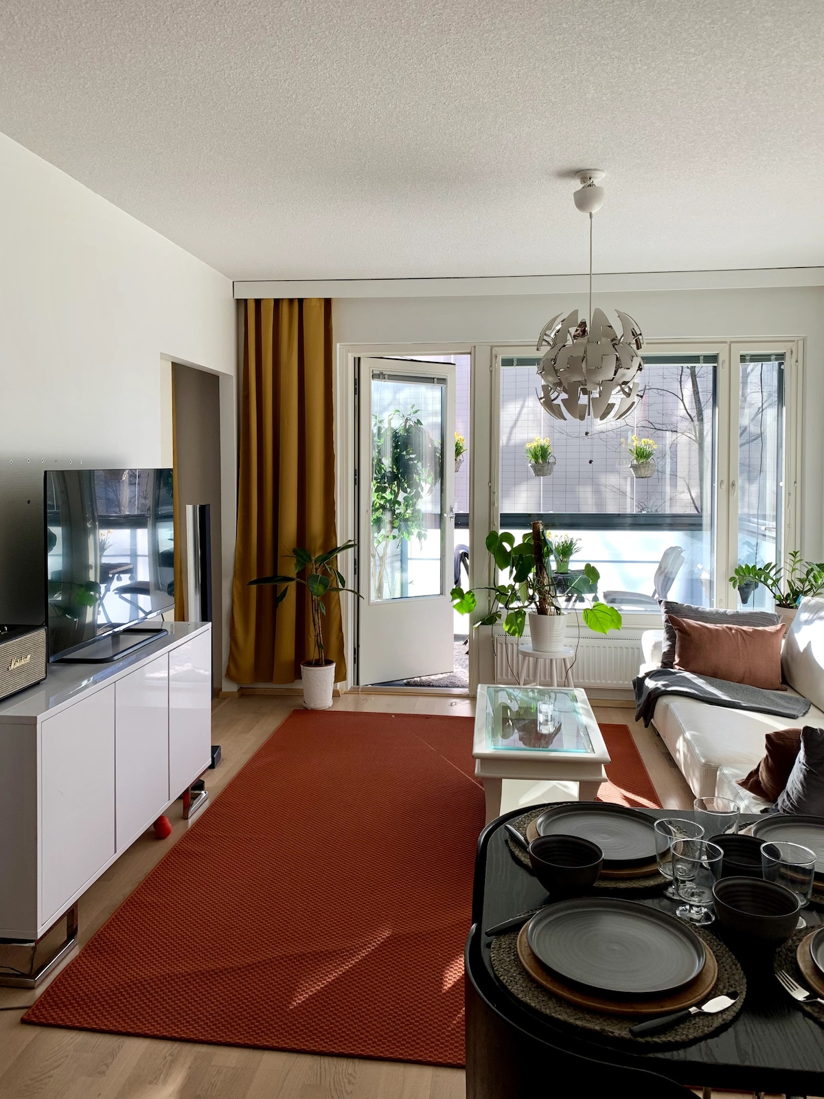 A modern living area features a comfortable sofa with decorative pillows and a glass coffee table. A television is positioned on a white cabinet, while a warm-toned rug complements the space. Large windows allow natural light to illuminate indoor plants placed nearby.
