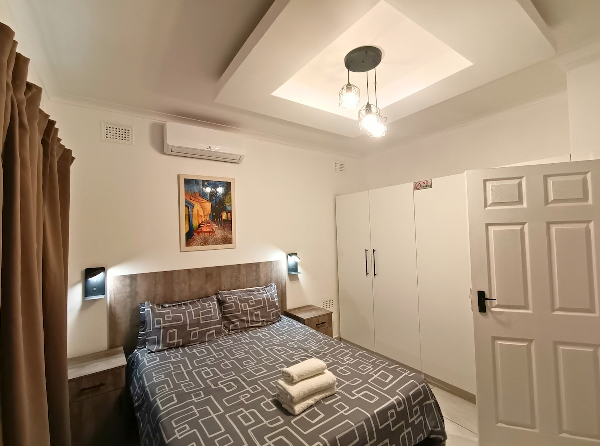 A well-lit bedroom features a comfortable bed with patterned bedding. Flanking the bed are two nightstands with wall-mounted reading lights. A wardrobe occupies one wall, and a piece of framed artwork adds a touch of color above the bed, contributing to a serene environment.