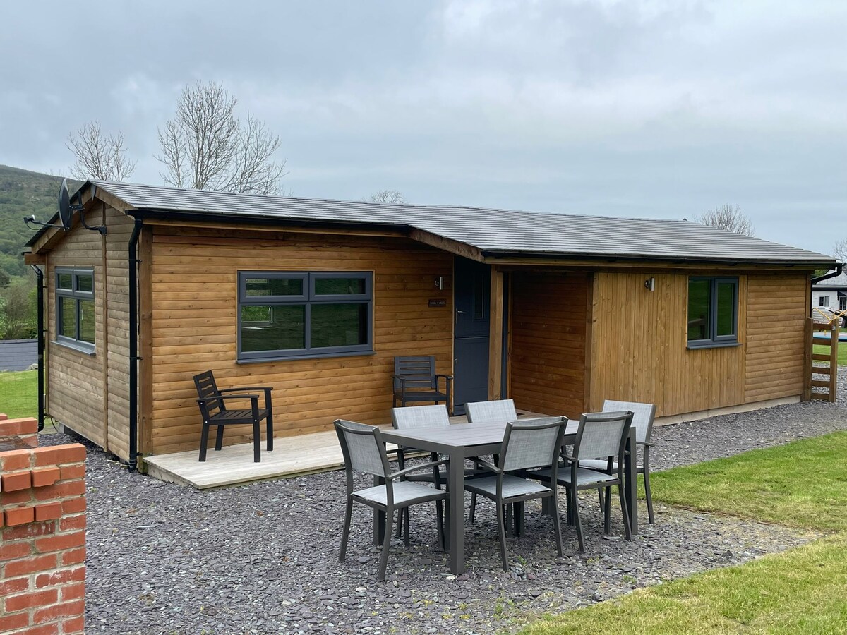 A wooden lodge features a covered entrance and is surrounded by a grassy area. Outdoor furniture, including a dining set with six chairs, is positioned on a patio. Large windows allow for natural light, and the building is set against a backdrop of rolling hills.