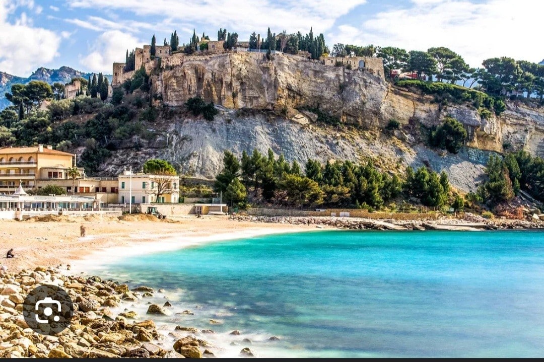 A clear view of a rocky coastline is framed by a sandy beach. The water displays various shades of turquoise, with gentle waves lapping against the shore. In the background, a fortified hillside offers a scenic contrast to the natural landscape.