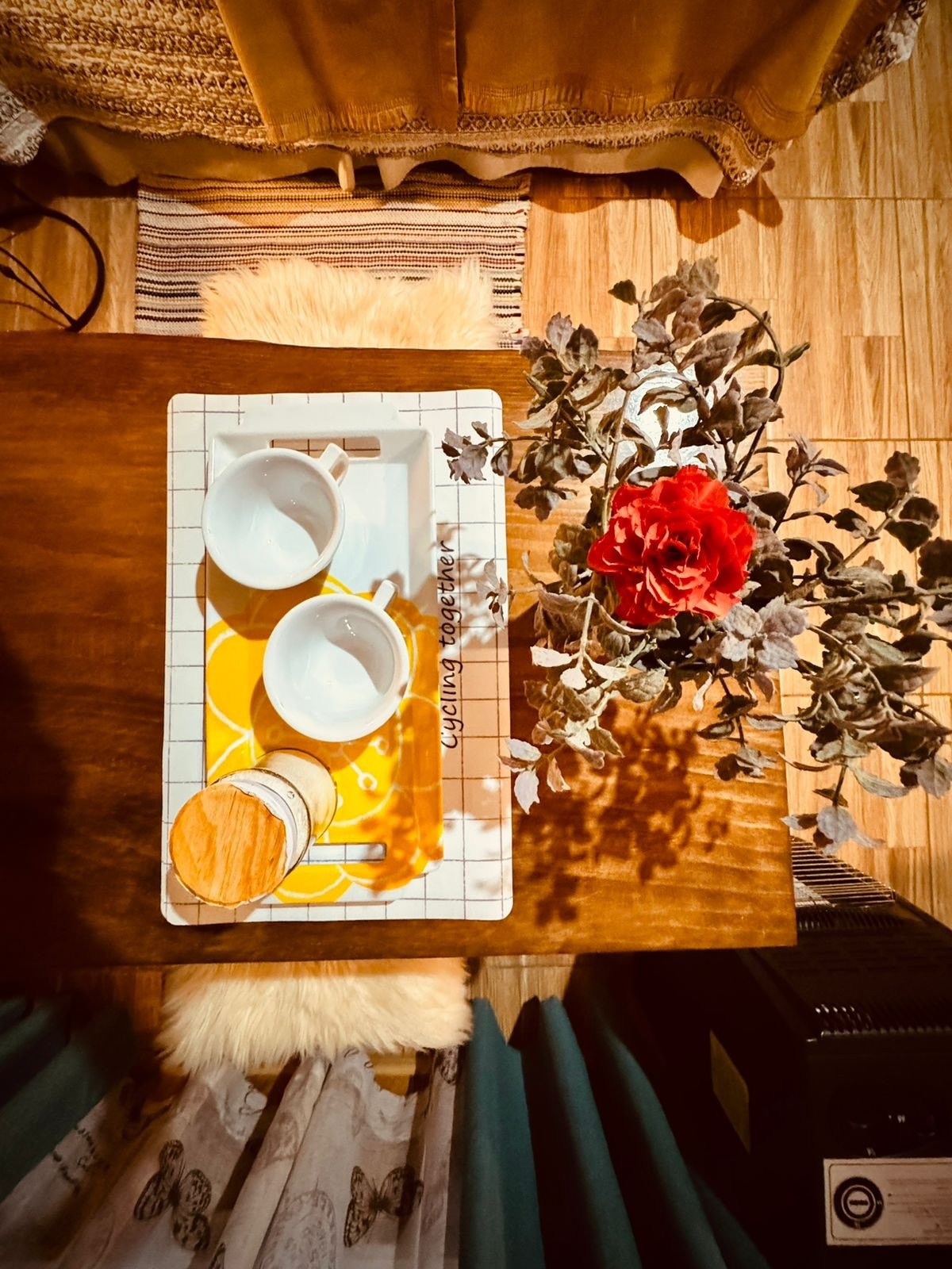 An overhead view of a wooden table is presented, featuring a decorative tray with two white cups and a stack of coasters. A small arrangement of foliage and a single red flower provides a pop of color, accentuating the warm, inviting setting.