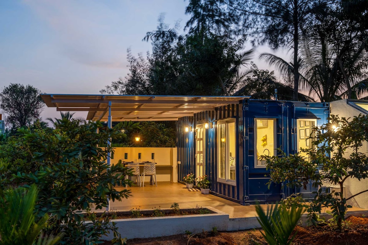 A charming container home is set within a lush landscape, featuring large sliding glass doors that offer views into the interior. An outdoor seating area is shaded by a roof, with a small dining table visible. Surrounding greenery creates a serene environment.