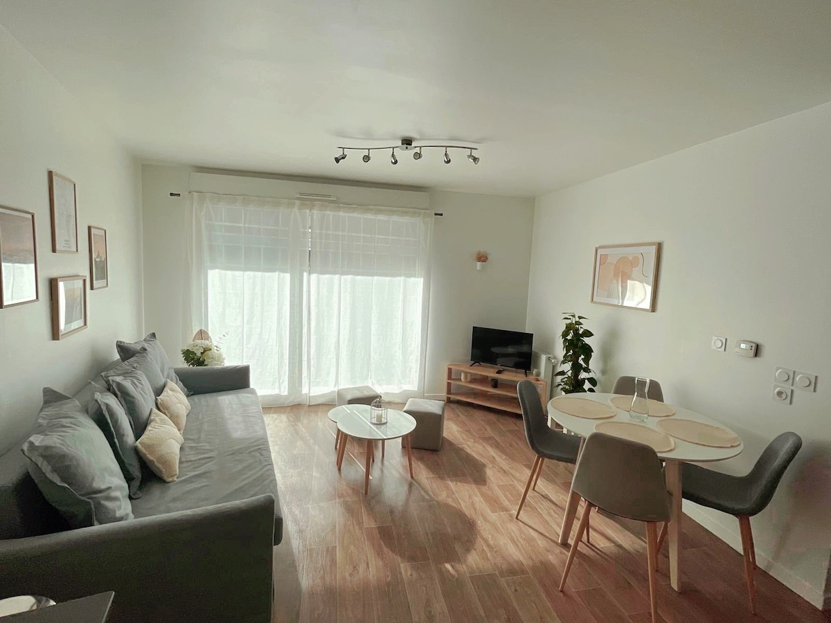 A bright living area is presented with a comfortable gray sofa and a modern coffee table. Light streams through large glass doors, illuminating the space. A round dining table with four chairs is positioned beside a small plant and a television unit, enhancing the functional layout.