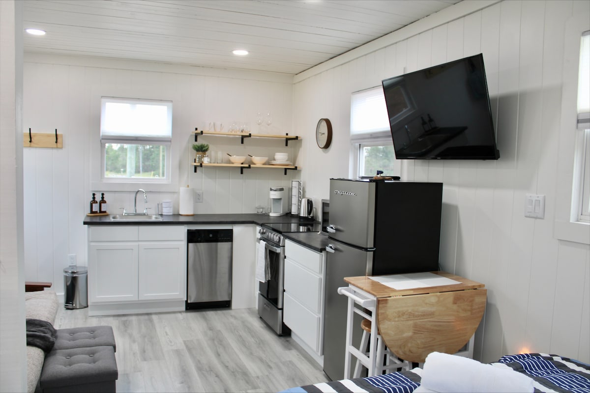 The kitchen area is designed with modern appliances, including a refrigerator and dishwasher. Open shelving displays dishware and utensils. A flat-screen TV is mounted on the wall, while a small table offers additional workspace. Natural light fills the space through two windows.
