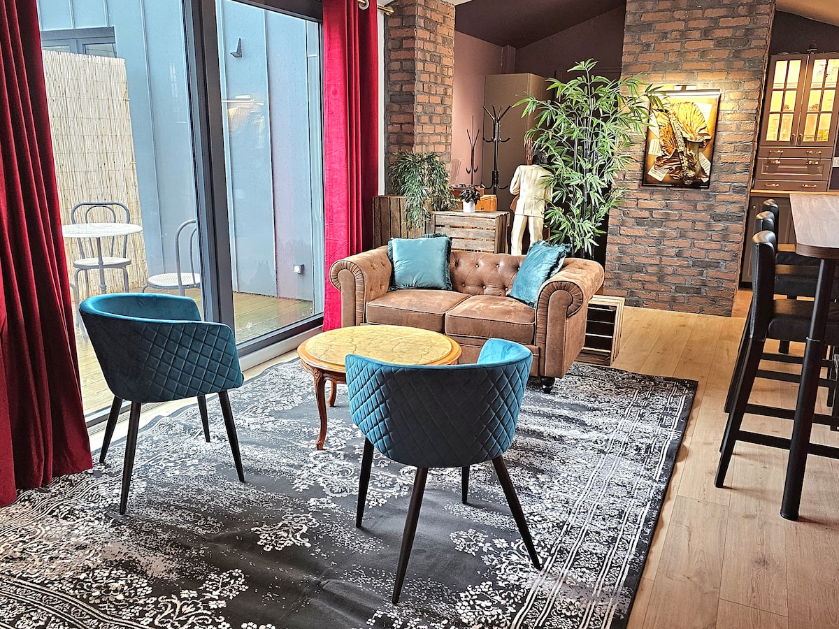 A stylish living room features a brown leather sofa paired with two teal chairs, creating a cozy seating area. A small round table rests on a patterned rug, with large glass doors providing natural light and a view of the outdoor space.