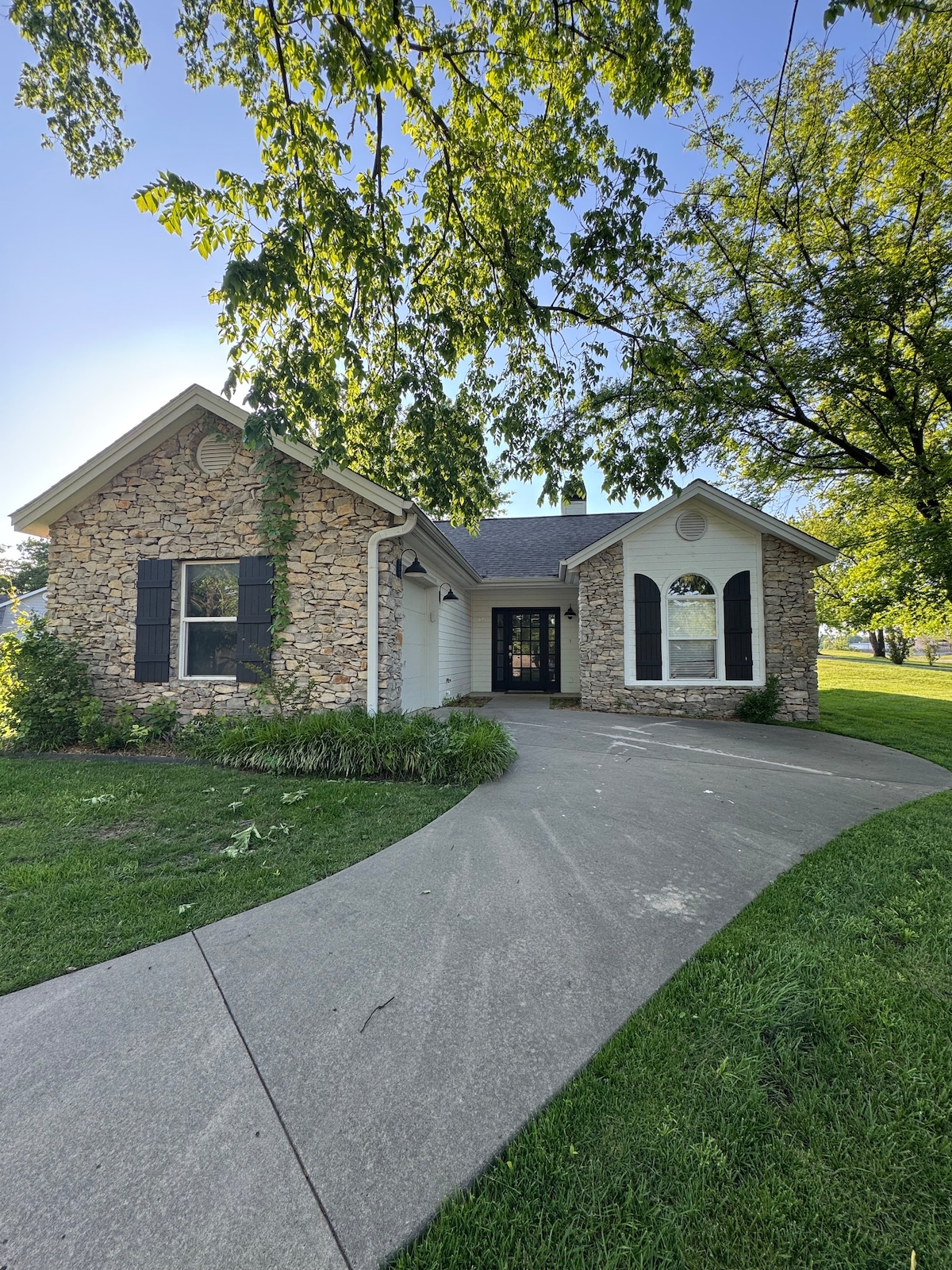 The exterior of the home features a charming stone façade, complemented by black shutters and decorative windows. A curved driveway leads to the entrance, flanked by well-maintained green grass and mature trees, creating a welcoming environment for guests.