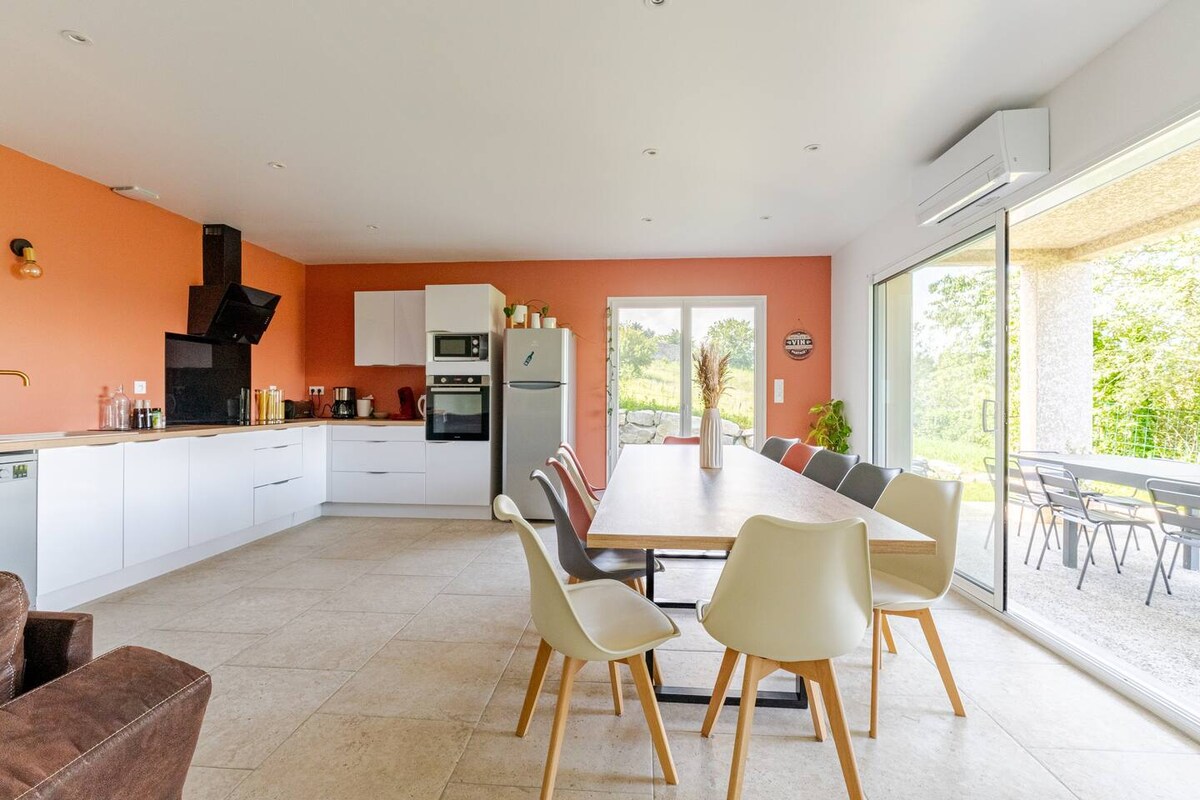 A spacious kitchen and dining area features modern appliances and a large table surrounded by six chairs in various colors. An ergonomic couch is present in the corner. Large glass doors open to an outdoor terrace with a view of the surrounding greenery.